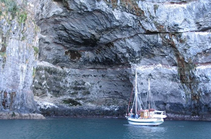 A sailing boat beneath pale volcanic sea cliffs with a cave at the waterline